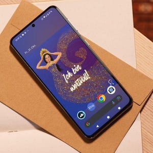 Könnte beinhalten: Ein schwarzes Smartphone zeigt eine Frau in einem goldenen Kleid und den deutschen Text "Ich bin wertvoll!" auf blauem und violettem Hintergrund. Das Telefon liegt auf einem braunen Umschlag und einem Notizbuch mit einem Stift.