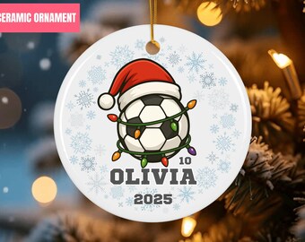 Op de afbeelding: Witte keramische kerstversiering met een voetbal met een kerstmuts en lichtsnoer. De versiering heeft de naam "OLIVIA" en het jaar "2025". De diameter van de versiering is ongeveer 7.6 cm.