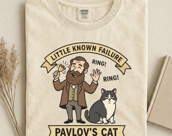 May include: Ivory-colored t-shirt featuring a cartoon illustration. The design includes a man ringing a bell, a cat, and text that reads "Little Known Failure" and "Pavlov's Cat". The shirt is made of a soft material.