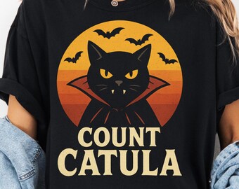 May include: Black t-shirt featuring a Halloween design. The graphic includes a black cat with yellow eyes and a vampire cape, set against a sunset background with bats. The text "COUNT CATULA" is below the cat.