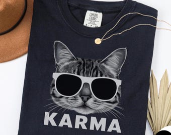 May include: Black t-shirt featuring a grayscale illustration of a cat wearing sunglasses, with the word "KARMA" in white block letters below. The shirt has a crew neck and a small tag on the upper back.