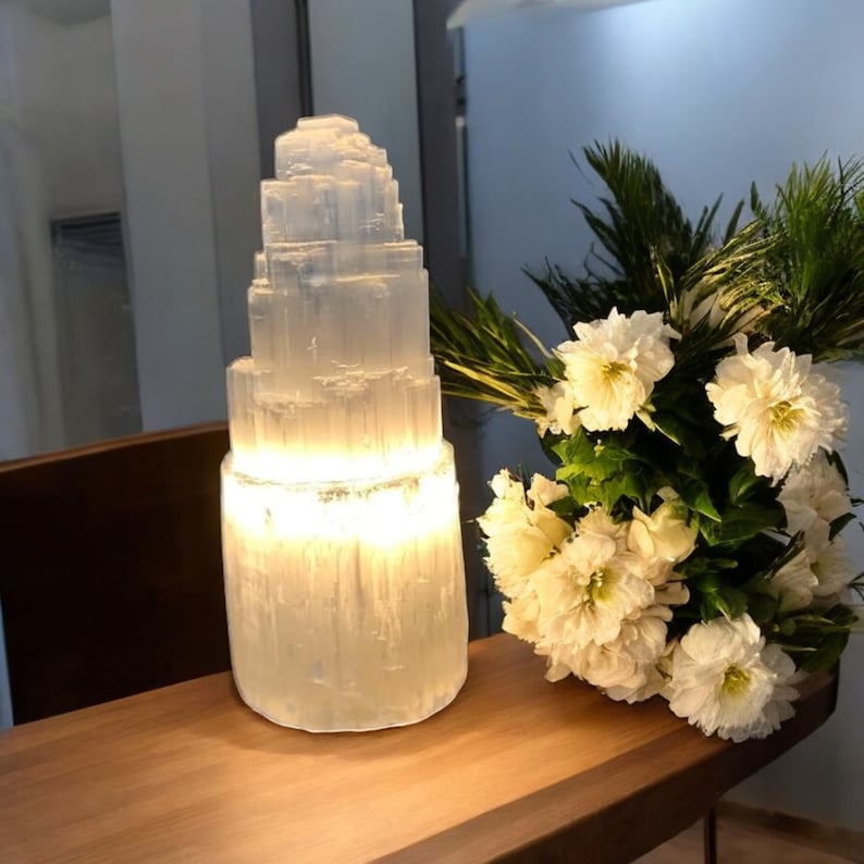 Peut inclure: Une lampe en cristal de s&eacute;l&eacute;nite blanche avec un design en couches, illumin&eacute;e de l'int&eacute;rieur, est pos&eacute;e sur une table en bois &agrave; c&ocirc;t&eacute; d'un bouquet de fleurs blanches.