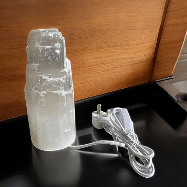 Peut inclure: Une lampe en cristal de s&eacute;l&eacute;nite blanche avec un cordon et une prise blancs. La lampe est pos&eacute;e sur une surface noire.