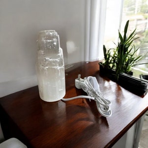 Peut inclure: Une lampe en cristal de s&eacute;l&eacute;nite blanche avec un cordon et une prise blancs. La lampe est pos&eacute;e sur une surface en bois brun fonc&eacute;.