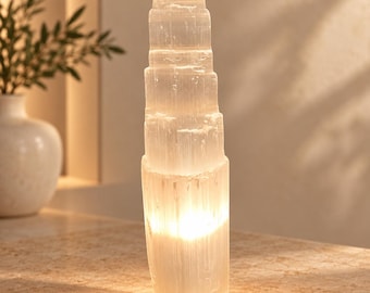 Könnte beinhalten: Eine hohe, zylindrische Selenit-Kristallturmlampe mit einer durchscheinenden, weißen, faserigen Textur. Der Stein hat ein gestapeltes, abgestuftes Aussehen mit einer hellen, warmen internen Lichtquelle, die von der Basis aus die vertikale Maserung beleuchtet.