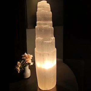 Peut inclure: Une lampe en cristal de s&eacute;l&eacute;nite blanche avec un design empil&eacute; et superpos&eacute;. La lampe est allum&eacute;e et diffuse une lueur chaude.