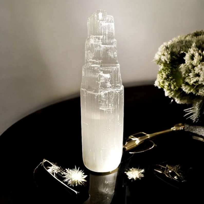 Peut inclure: Une tour de cristal de s&eacute;l&eacute;nite blanche avec un design en couches superpos&eacute;es. Le cristal est pos&eacute; sur une surface noire avec un objet en m&eacute;tal dor&eacute; et quelques petits objets en forme d'&eacute;toile argent&eacute;s.