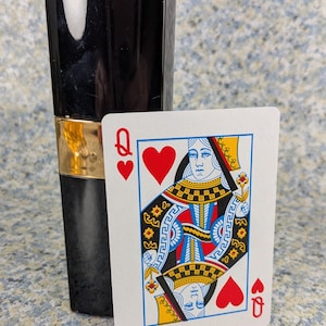 May include: A black rectangular perfume bottle with a gold band is next to a playing card. The card is the Queen of Hearts, featuring a portrait of a queen, red hearts, and the letter "Q".