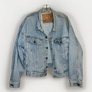May include: Light blue denim jacket with a classic design. Features a button-up front, two chest pockets, and two side pockets. The jacket has a faded look and a brown label on the back. A timeless piece for any wardrobe.