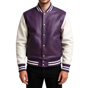 Puede incluir: Chaqueta universitaria morada y crema. La chaqueta tiene un cuerpo de cuero morado con mangas crema. El cuello, los puños y la cintura tienen rayas blancas. Tiene cierre de botones y dos bolsillos delanteros.