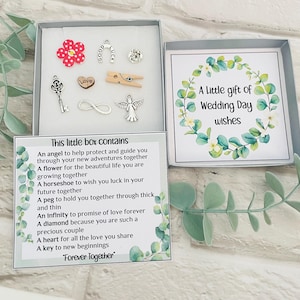 May include: A small white box with a floral design and the text "A Little gift of Wedding Day wishes". The box contains a variety of small charms, including a flower, a horseshoe, a peg, an infinity symbol, a heart, and a key. The box also includes a card with the text "This little box contains..." and a list of what each charm represents.