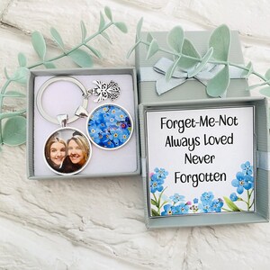 Puede incluir: Un llavero plateado con un dije de foto y un dije de no me olvides. El dije de foto presenta una imagen de dos mujeres. El llavero viene en una caja gris con un diseño de no me olvides y el texto "Forget-Me-Not Always Loved Never Forgotten".
