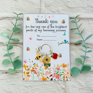 Könnte beinhalten: Ein Schlüsselanhänger mit Bienen-, Sonnenblumen- und Apfelanhängern, befestigt an einer Karte mit dem Text "Thank you for bee-ing one of the brightest parts of my learning journey." Die Karte hat ein Blumenmuster und Platz für "An" und "Von".