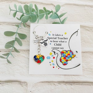 May include: A silver keychain with a heart-shaped charm featuring a puzzle piece design in red, yellow, blue, and green. The keychain also includes two smaller charms, one with the word "Teacher" and the other with the word "Thank You". The keychain is displayed on a card with the text "It takes a Special Teacher to hear what a Child cannot say".