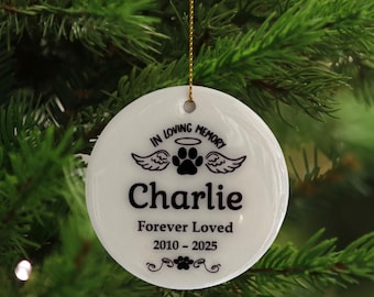 May include: A white, round Christmas ornament with the text "IN LOVING MEMORY" above a paw print and angel wings design. The name "Charlie" is centered below, with "Forever Loved" and the years 2010-2025 below that. A gold string is attached.