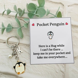 May include: A silver keyring with a white pearl charm and a black and white penguin charm. The keyring also has a silver letter K charm. A white card with red hearts and the text "Pocket Penguin" is attached to the keyring. The card has a black and white penguin illustration and the text "Here is a Hug while I can't be there... keep me in your pocket and take me everywhere..."