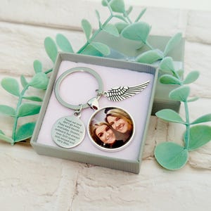Könnte beinhalten: Ein silberner Schlüsselanhänger mit einem runden Fotoanhänger und einem Flügel-Charm, präsentiert in einer grauen Geschenkbox. Der Fotoanhänger zeigt zwei lächelnde Gesichter. Ein runder Charm mit graviertem Text ist ebenfalls angebracht. Grüne Zweige umgeben die Box.