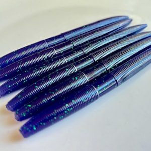 May include: A close-up of a pack of dark blue and green fishing lures. The lures have a ribbed texture and a pointed end.