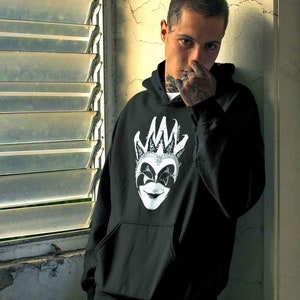 May include: Black hoodie featuring a white and gray graphic of a jester mask. The design includes a crown and a smiling face. The hoodie has a front pocket and a drawstring hood. The person in the image is wearing the hoodie.