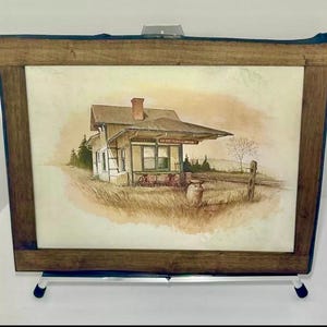Könnte beinhalten: Ein gerahmtes Aquarellgemälde eines alten Bahnhofs. Das Kunstwerk zeigt ein beiges und weißes Gebäude mit einem roten Schornstein vor dem Hintergrund eines Feldes und Bäumen. Der Rahmen besteht aus dunkelbraunem Holz.