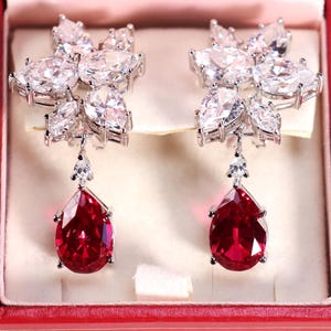 May include: A pair of silver-toned earrings featuring a cluster of clear, pear-shaped and marquise-cut stones at the top, with a single, large, teardrop-shaped red stone suspended from each, all set in a metallic frame.