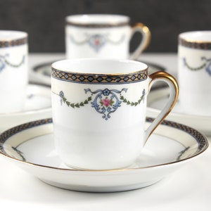 May include: Set of six white porcelain teacups and saucers with gold trim and floral designs. The cups have a delicate floral pattern in blue and pink with gold accents. The saucers have a gold rim and a matching floral design.