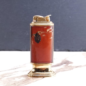May include: A rectangular table lighter with a polished gold-tone metal base and top mechanism. The central body features a deep amber-colored panel with a dark brown pinecone graphic and subtle branch detailing on the upper left side.