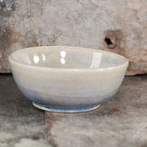 May include: A small, white ceramic bowl with a blue glaze on the bottom. The bowl is sitting on a gray, textured surface.
