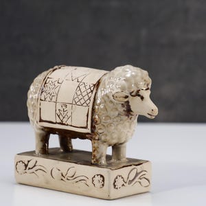 May include: A ceramic sheep figurine in an off-white glaze with brown accents. The sheep features a textured wool coat and a blanket on its back with a geometric pattern. It stands on a rectangular base decorated with embossed floral motifs.