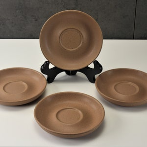 May include: Set of four brown ceramic saucers with a slightly raised center. The saucers are arranged in a triangular pattern on a white surface.