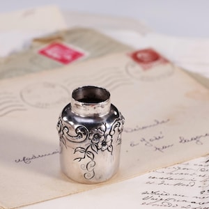 May include: A small, silver inkwell with floral and scrollwork designs. The inkwell sits on a vintage letter with handwritten script. The inkwell has a cylindrical shape with a wide opening and a decorative band around the middle.