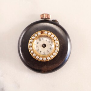May include: A small, round, antique pocket watch with a dark brown casing. The watch face is white with black numerals and hands, surrounded by a gold bezel with small, white pearls. A copper-colored winding stem is visible.