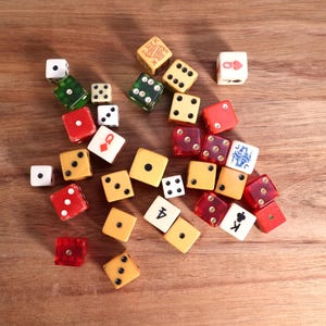 May include: A collection of dice in various colors, including red, yellow, green, and white, scattered on a wooden surface. Some dice feature pips, while others display playing card symbols. The dice are of different sizes and shapes.