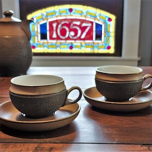 Puede incluir: Dos tazas de té de cerámica marrón con interiores blancos y platillos sobre una mesa de madera. Las tazas de té tienen un acabado texturizado y moteado. Las tazas de té están frente a una ventana con un panel de vitral que presenta el número 1657.