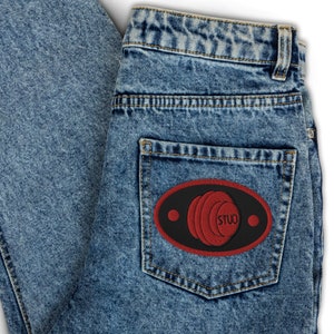 Può includere: Jeans di denim blu chiaro con una tasca posteriore con una toppa ricamata. La toppa è nera con un design ovale rosso e la parola "STUD" in bianco. I jeans hanno un classico design a cinque tasche.