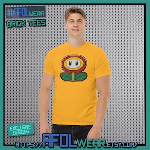 Può includere: Una maglietta gialla con un disegno di fiore a cartoni animati nei colori rosso, giallo e verde. La maglietta mostra il testo "#AFOLWEAR BRICK TEES" e "EXCLUSIVE DESIGNS". L'URL del negozio Etsy è visibile.