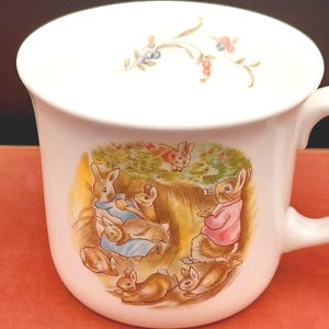 May include: A white ceramic mug featuring a colorful illustration of several rabbits in a garden setting. One rabbit wears a pink jacket, while others are depicted in various poses. A small floral sprig is painted on the upper interior rim.