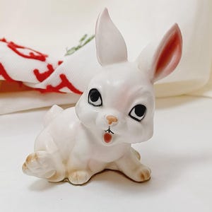 May include: A white ceramic bunny figurine with pink ears and black eyes. The bunny is sitting on its hind legs with its front paws outstretched.