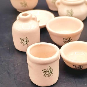 May include: A collection of miniature cream-colored ceramic kitchenware. The set includes bowls, a pitcher, a lidded pot, and a small jar, each adorned with a green leaf design. The items are arranged on a dark surface.