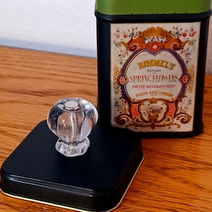 May include: A black metal tin with a green rim and a label reading Rimmel's Extract of Spring Flowers for the Handkerchief Paris and London. The tin has a separate black lid featuring a clear, faceted, glass-like knob handle on top.
