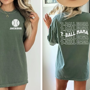 May include: An olive green short-sleeve t-shirt featuring a white baseball graphic and the name JACKSON on the front left chest. The back of the shirt displays the repeated white text T-BALL MAMA in a stacked, wavy, retro-style font design.