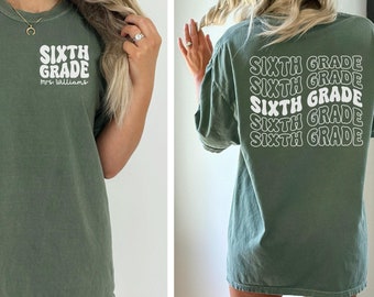 May include: An olive green t-shirt featuring the text "SIXTH GRADE" in a white, retro font on the front and back. The front also includes "Mrs. Williams" below the main text. The back of the shirt repeats the text in a stacked design.