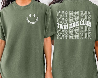 Puede incluir: Camiseta verde oliva con la inscripción blanca "TWIN MOM CLUB" en la parte delantera y trasera. La parte delantera presenta un diseño de carita sonriente con las palabras "TWIN MOM CLUB" en un círculo. La parte trasera tiene las palabras "TWIN MOM CLUB" repetidas.