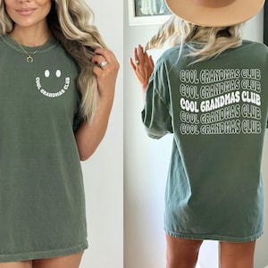 May include: Olive green t-shirt with the text "COOL GRANDMAS CLUB" in white on the back. The front of the shirt features a smiling face graphic with the text "COOL GRANDMAS CLUB" in a circular design.