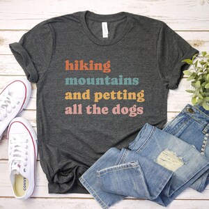 May include: A dark gray t-shirt with the text "hiking mountains and petting all the dogs" in colorful letters. The shirt is laid flat on a white wooden surface with a pair of white sneakers and blue jeans.