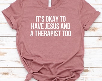 Puede incluir: Una camiseta de color malva con el texto "It's okay to have Jesus and a therapist too" impreso en blanco.