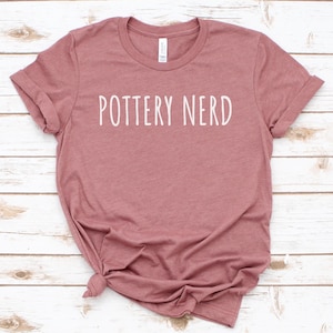 May include: A dusty rose-colored t-shirt with the words "POTTERY NERD" printed in white, centered on the front. The shirt has short sleeves and a crew neck. The fabric is slightly wrinkled, and the shirt is knotted at the bottom.