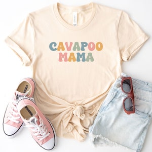 May include: A light beige t-shirt with the text "CAVAPOO MAMA" in colorful, retro-style letters. The shirt is paired with light blue denim shorts and pink Converse sneakers.