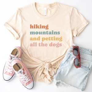 May include: A beige t-shirt with the text "hiking mountains and petting all the dogs" in colorful retro font. The shirt is paired with light blue denim shorts and pink sneakers.