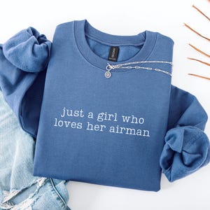 Könnte beinhalten: Ein blaues Sweatshirt mit Rundhalsausschnitt und dem weiß gestickten Text "just a girl who loves her airman" auf der Brust. Das Kleidungsstück wird mit einer abgenutzten blauen Jeans und einer silberfarbenen Kette mit kleinem Anhänger kombiniert.